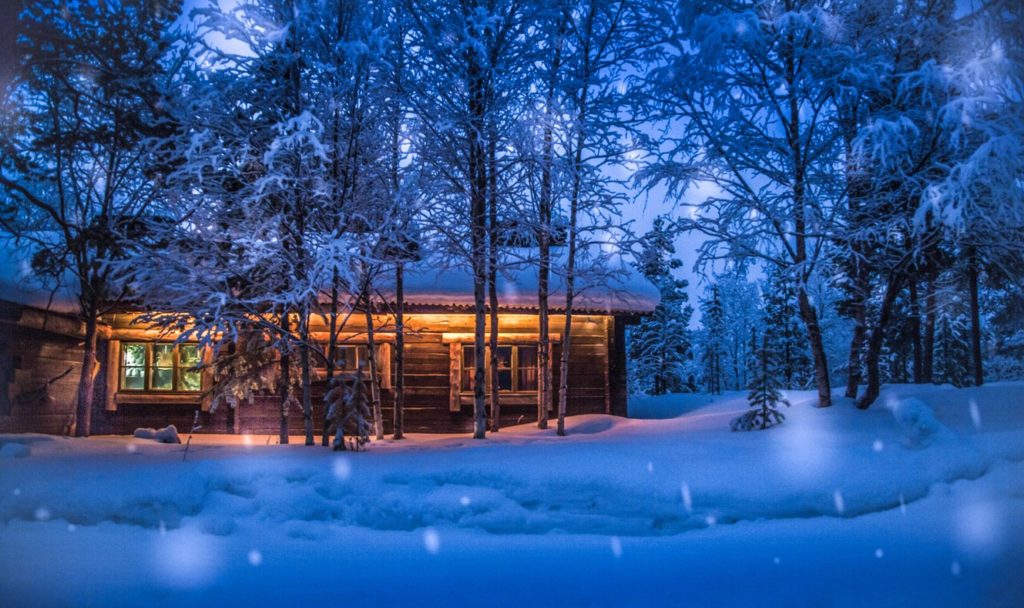 A cabin lit up on the outside in the evening in snowy woods with snow falling.