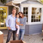A mother, father and their child stand outside their new home waving.